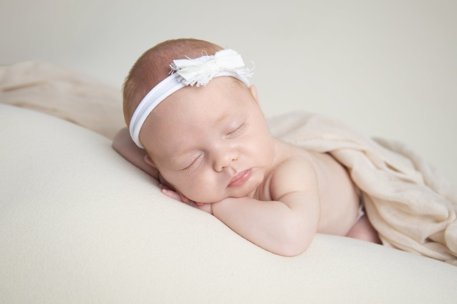 A sleeping newborn baby wearing a white bow headband on a soft cream surface